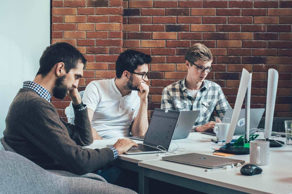 3 men are sitting, looking at their laptops